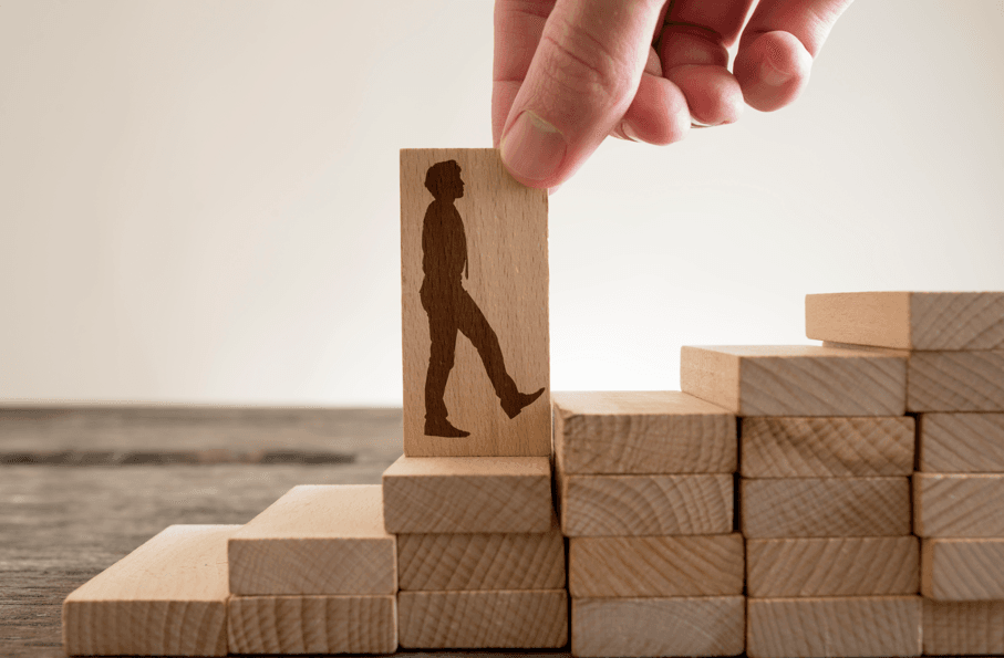Avaliação Insatisfatória no Estágio Probatório: homem segurando com os dedos o dominó de madeira com forma de empresário subindo escadas para subir a escada da carreira.