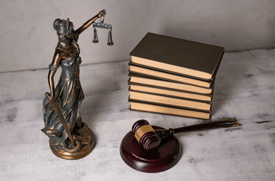 Lei de improbidade administrativa: estátua da deusa da justiça segurando balança, ao lado de pilha de livros jurídicos e martelo de juiz sobre a mesa.